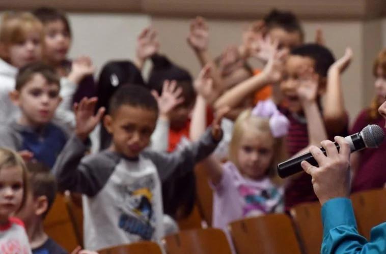 A room of small children with hands raised, responding to Michael Polzin, off-camera at right, holding a microphone.