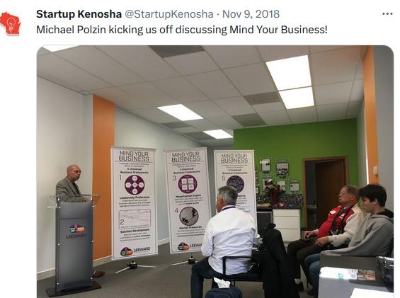 Michael Polzin at a podium delivering the Mind Your Business workshop, banners visible behind him.
